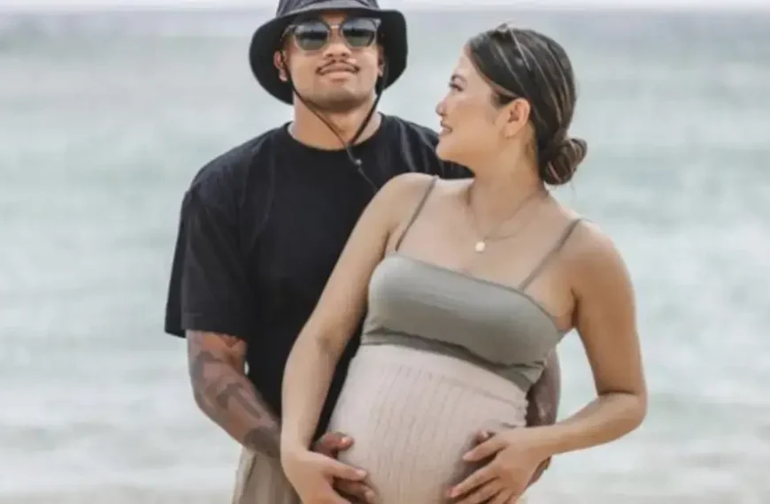 couple maternity poses hand on her belly while standing in front of the sea