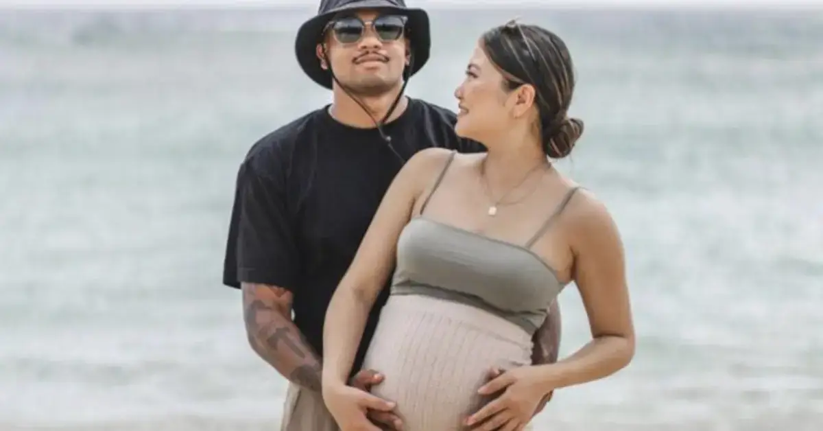 couple maternity poses hand on her belly while standing in front of the sea